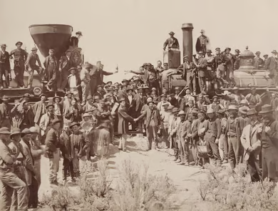 Sepia-toned photograph of crews from the Union Pacific and Central Pacific Railroads gathered between two locomotives at Promontory Summit, Utah, marking the joining of the transcontinental railroad on May 10, 1869.